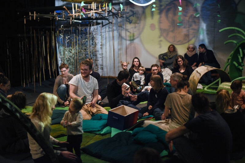 Kleinkinder sitzen mit den Schauspieler:innen und ihren Eltern auf Kissen und zwischen verschiedenen Klangkörpern 