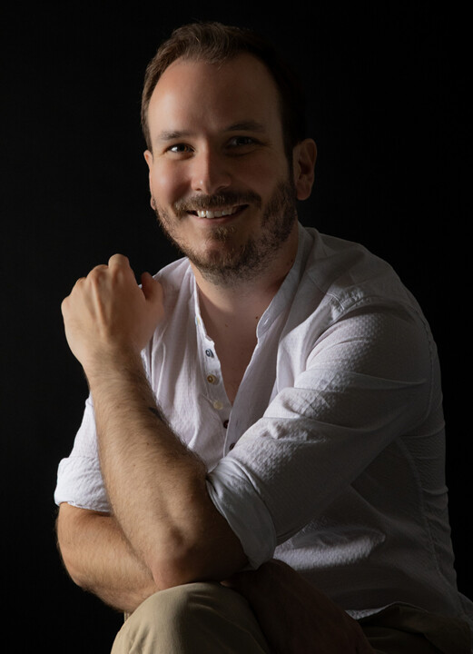 Mann mit hochgekrempeltem weißem Hemd sitzt vor schwarzem Hintergrund, Arm angewinkelt und Hand zur Faust geballt.