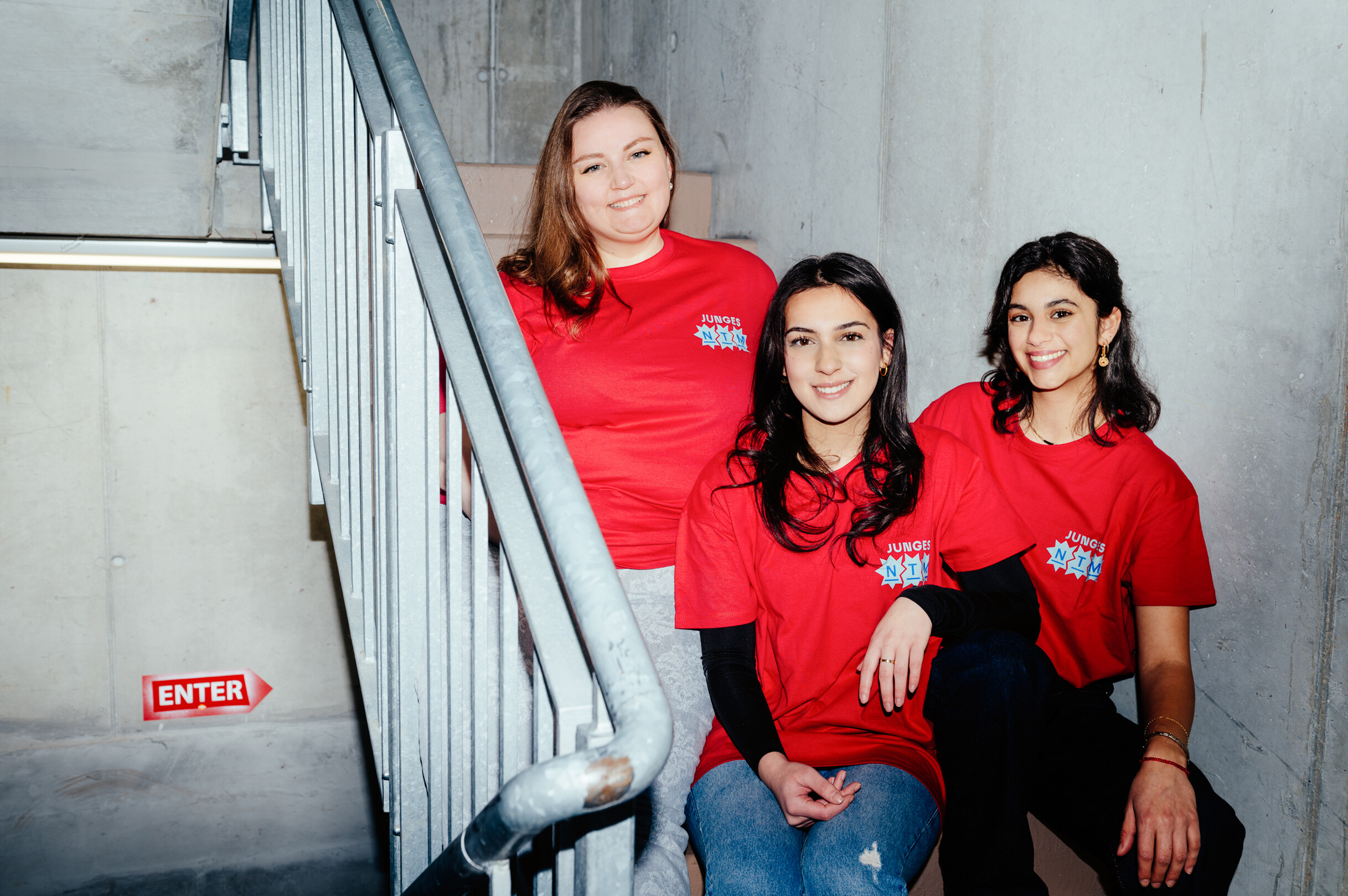 Drei junge Frauen in roten T-Shirts sitzen auf einer Treppe und lachen in die Kamera.