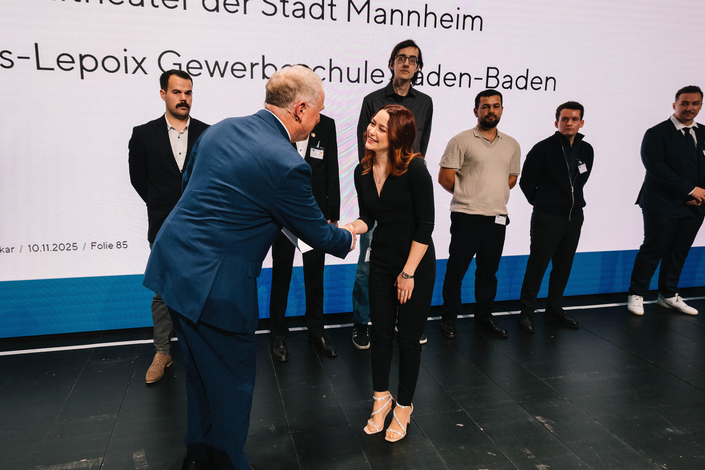 Mann im blauen Anzug schüttelt einer Frau in schwarzer Kleidung auf einer Bühne die Hand, mehrere Personen stehen im Hintergrund vor einer Präsentationswand.