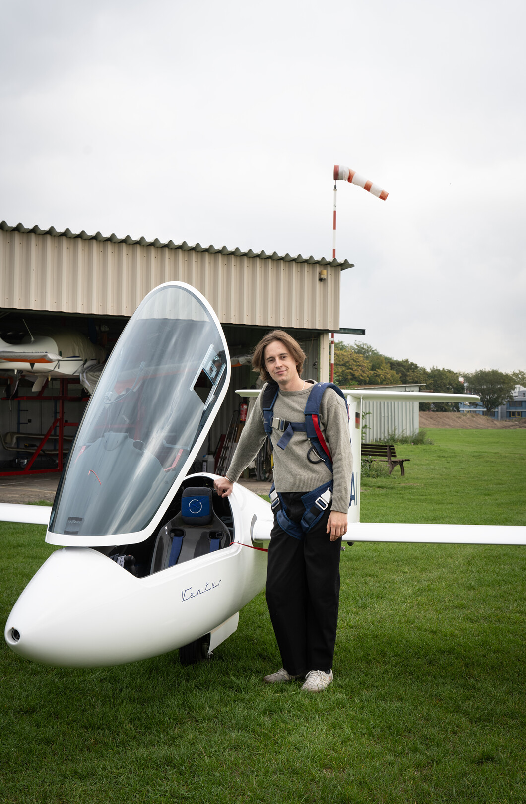 Man sieht einen jungen Mann mit mittellangen dunkelblonden Haaren, der auf einem Flugplatz neben einem Segelflugzeug steht.