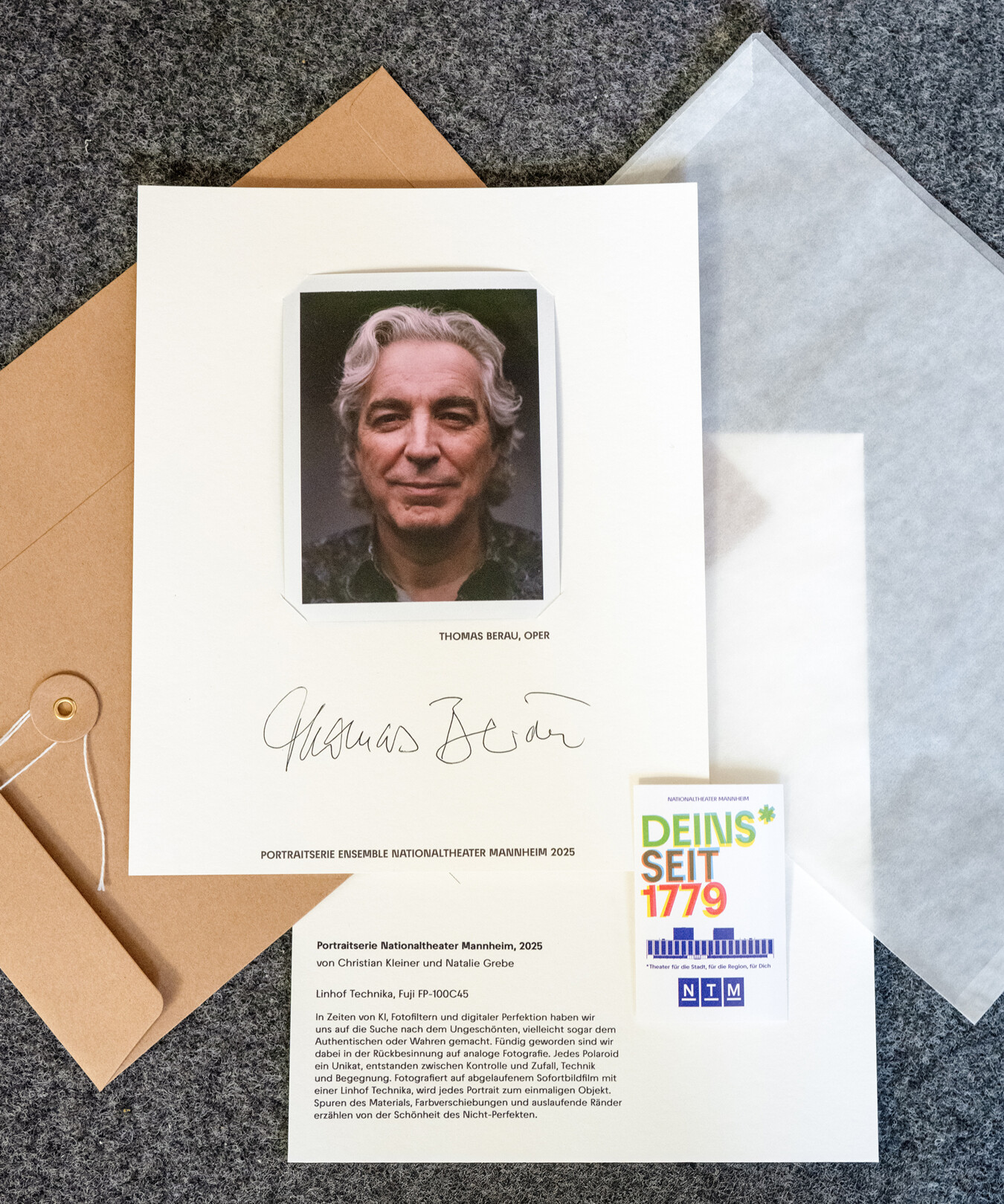 Polaroid-Fotografie einer Person mit grauen Haaren auf einem weißen Blatt mit Unterschrift und Text, daneben eine Karte mit der Aufschrift 'DEINS* SEIT 1779'.