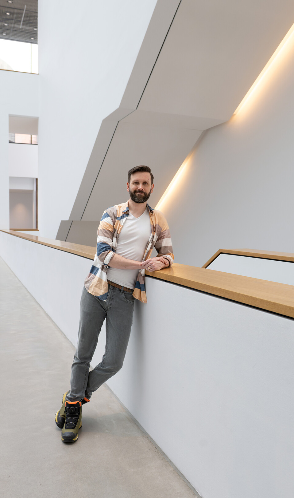 Christopher steht an ein weißes Geländer gelehnt, hat die Hände ineinander verschränkt und schaut lächelnd in die Kamera. Er trägt ein bunt kariertes Hemd und eine schwarze Hose.