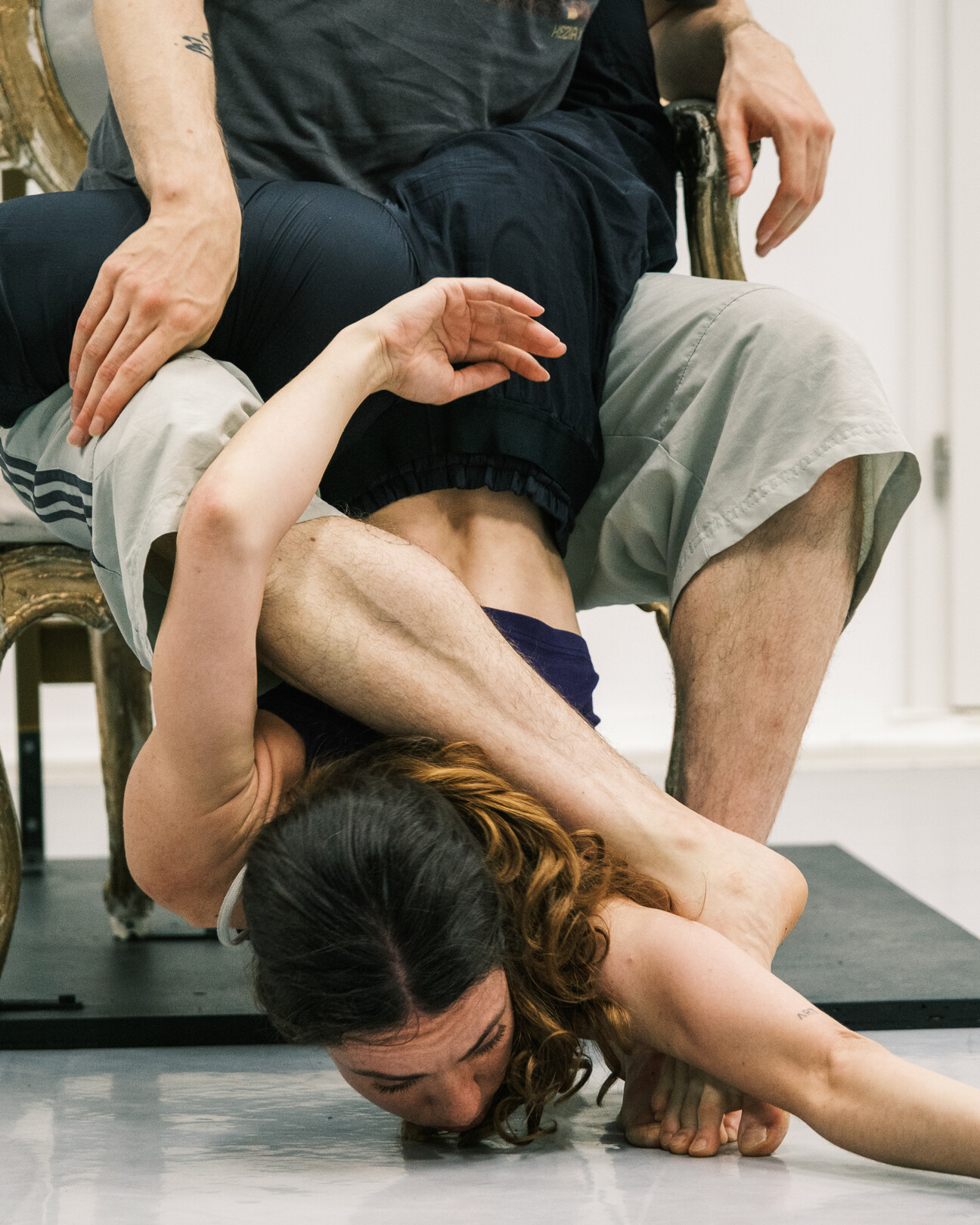 Person in schwarzer Sportkleidung liegt rückwärts über den Beinen einer sitzenden Person in grauer Hose und schwarzem T-Shirt, beide auf Matte.